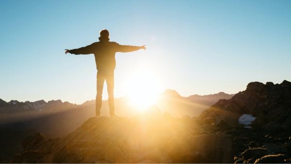 Person mit ausgestreckten Armen auf einem Berggipfel mit Blick auf die aufgehende Sonne